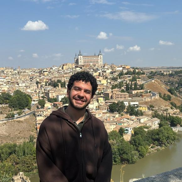 A male is sitting on a fence. There is lake and buildings behind him