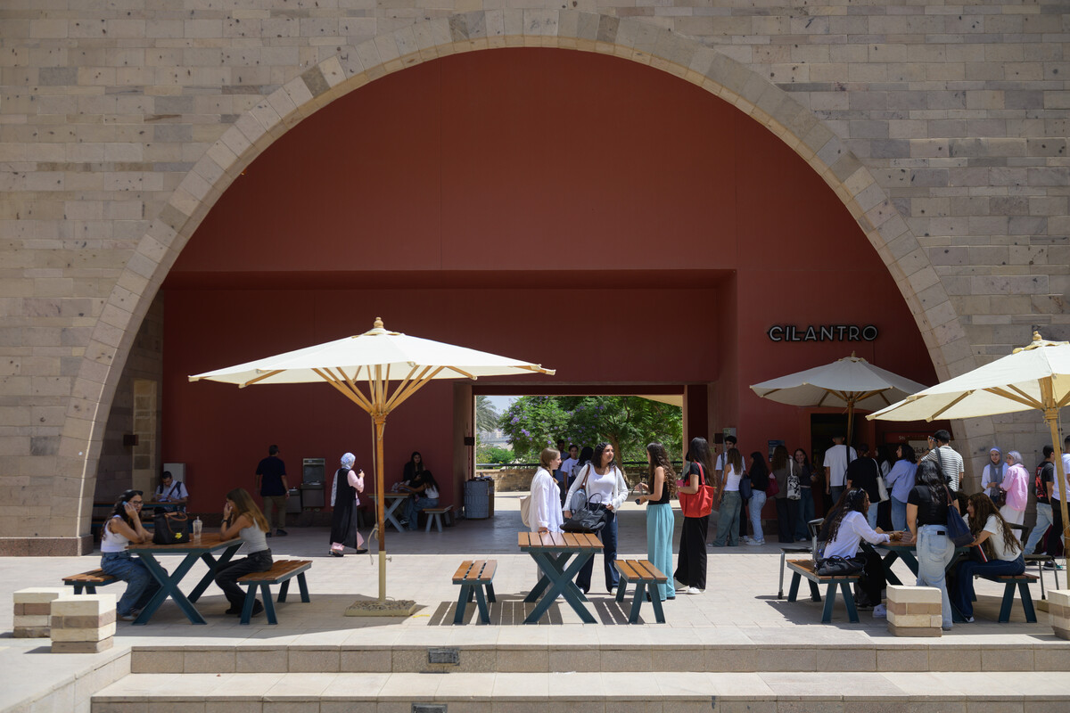 People sitting on benches and standing outdoors under shades talking and eating. There is a queue in front of a food outlet. Text: Cilantro 