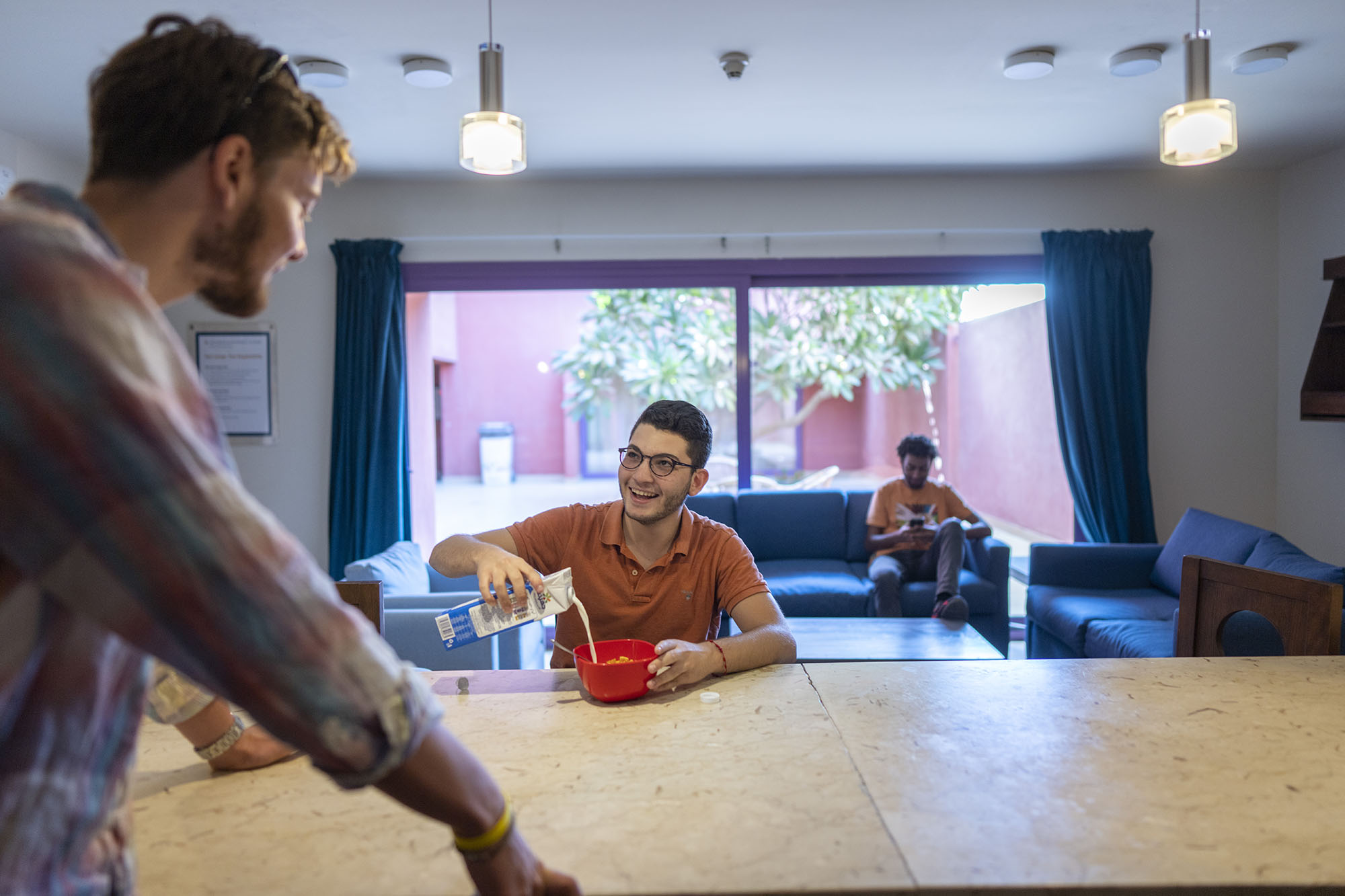 Two male students are talking together, one is standing up and the other is pouring milk from a carton into a bowl. Another male student is sitting in the background reading on a sofa
