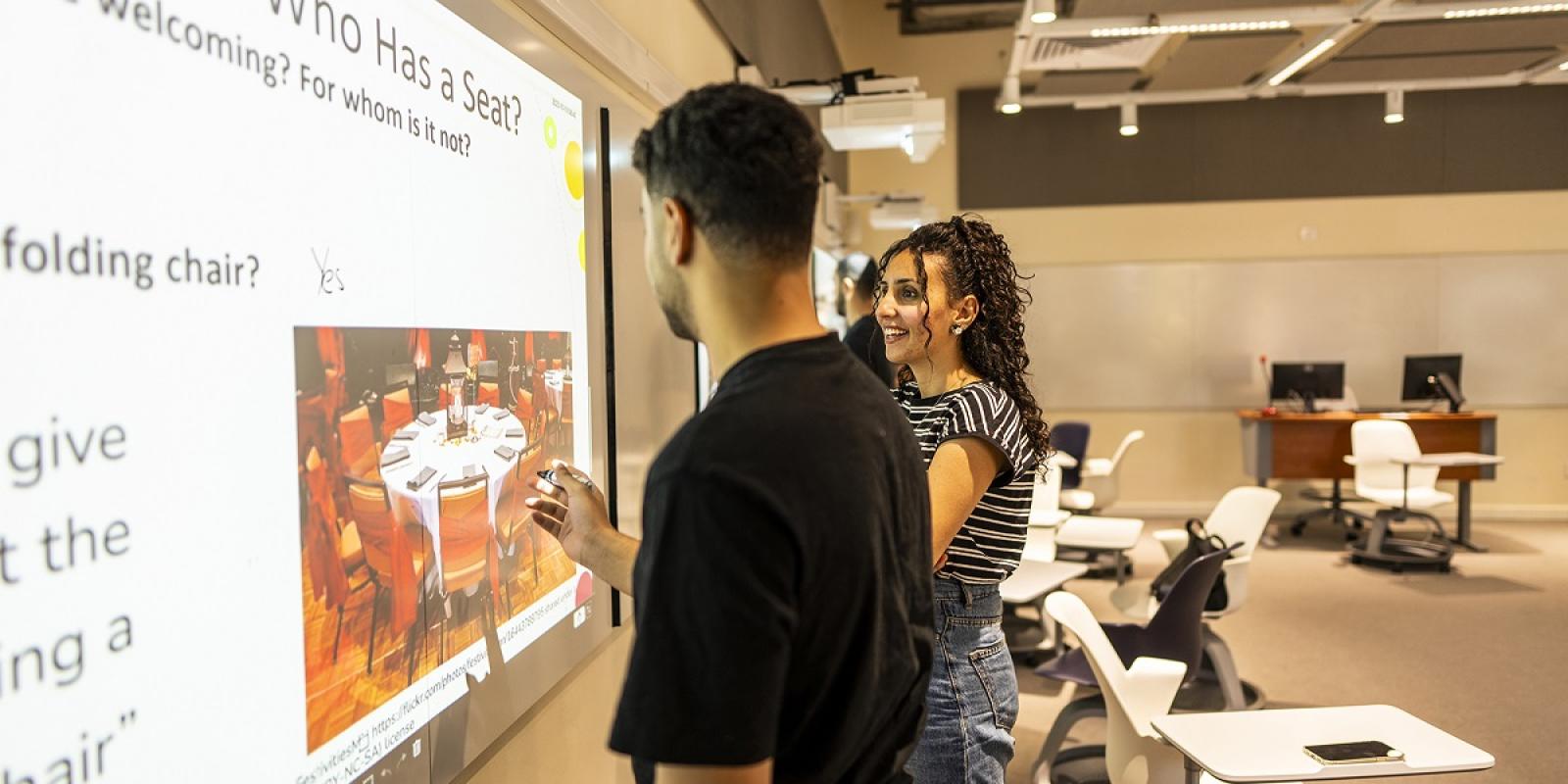 Males and a female standing in front of screens with an English text. The males are writing on the screens