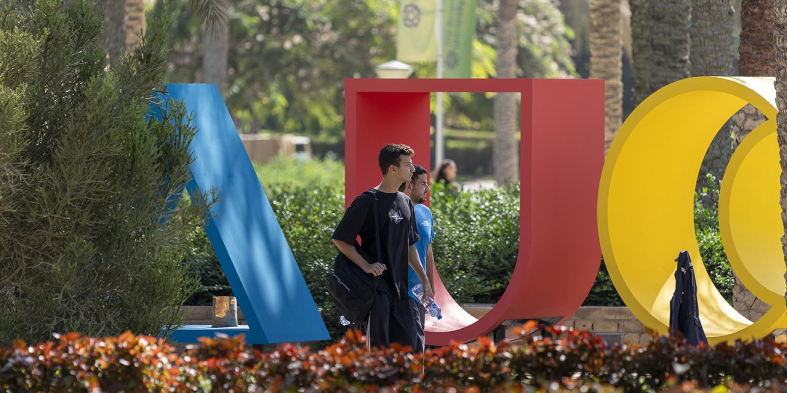 Two males are walking beside big A U C letters in a garden
