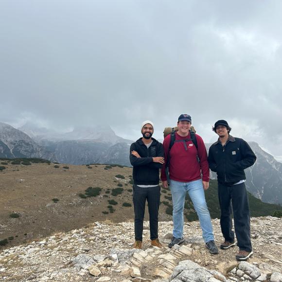 Three males are standing on a mountain top and smiling. There's fog around them