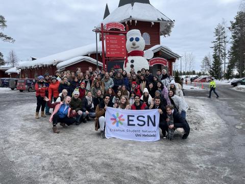 Male and females standing and sitting in the snow in front of a windmill. They are holding a sign with the text "ESN Erasmus Student Network. Uni Helsinki"
