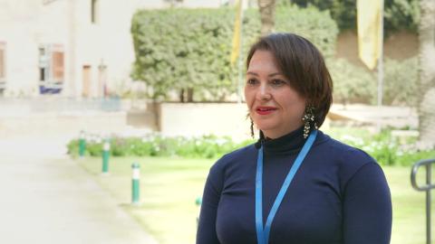 A woman is smiling. She is standing in a garden.