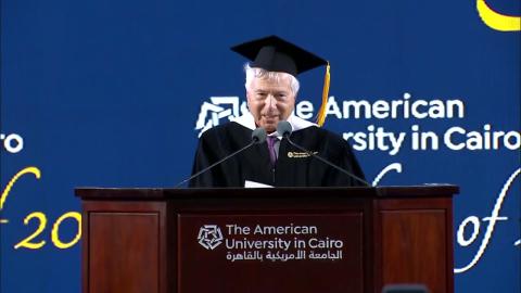A man is wearing a cap and gown. He is standing at a podium and talking into a microphone. Text: The American University in Cairo