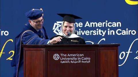 A woman is wearing a cap and gown. She is standing at a podium and talking into a microphone and a man is helping her to fix the microphone. Text: The American University in Cairo