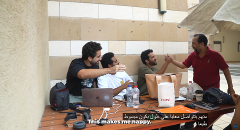 A male is shaking hands with three males sitting down at a desk. They are laughing