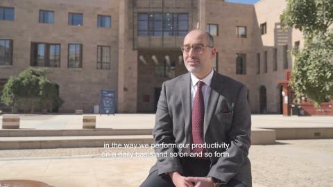 A man is sitting in an open space, there are building around him. He is wearing a suit and glasses